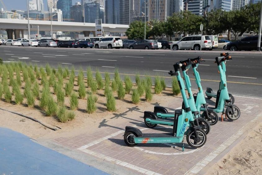 E-Roller geparkt vor einem Wolkenkratzer in Dubai mit grünen Pflanzen auf der Straßenbegrenzung.