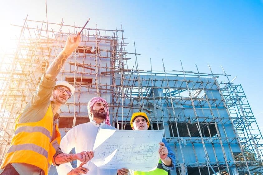 Dubai Hausbau, eingerüstetes Gebäude im Vordergrund, Emirati-Bürger, der einen Bauplan hält, berät sich mit zwei Bauingenieuren.
