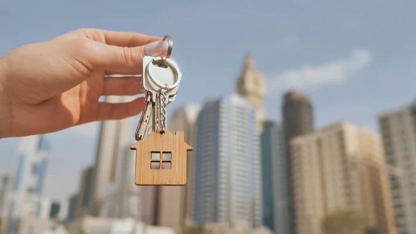 Ein Schlüssel mit einem Holz-Hausschlüsselanhänger, verschwommene Wolkenkratzer von Dubai im Hintergrund.