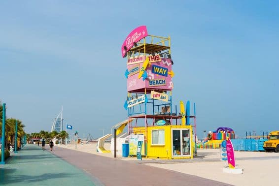 Dubai Kite Beach Willkommensstruktur, Schilder, Burj Al Arab im Hintergrund, Vordergrund mit Laufstrecke und Sandstrand.