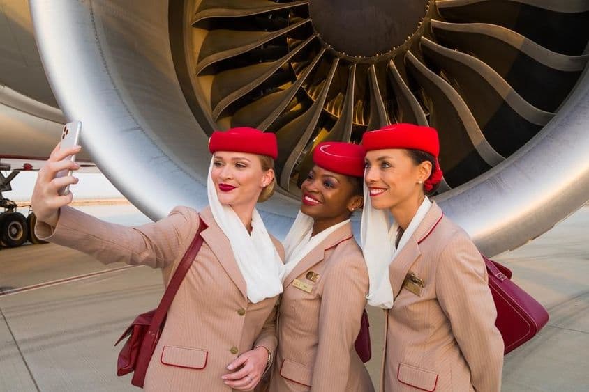 Drei Emirates-Flugbegleiter machen ein Selfie vor einem Emirates-Flugzeugmotor.