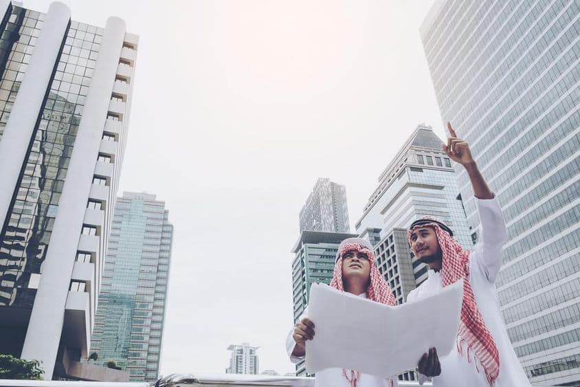 Zwei arabische Designer mit einem Bauplan blicken in die Ferne, Wolkenkratzer hinter ihnen.