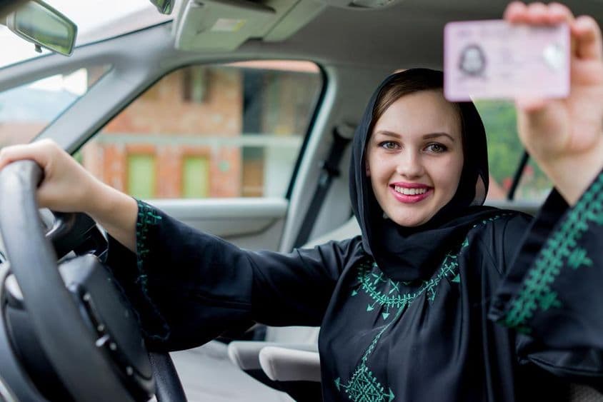 Muslimische Frau am Steuer eines Autos, hält einen Führerschein, strahlt vor Freude.