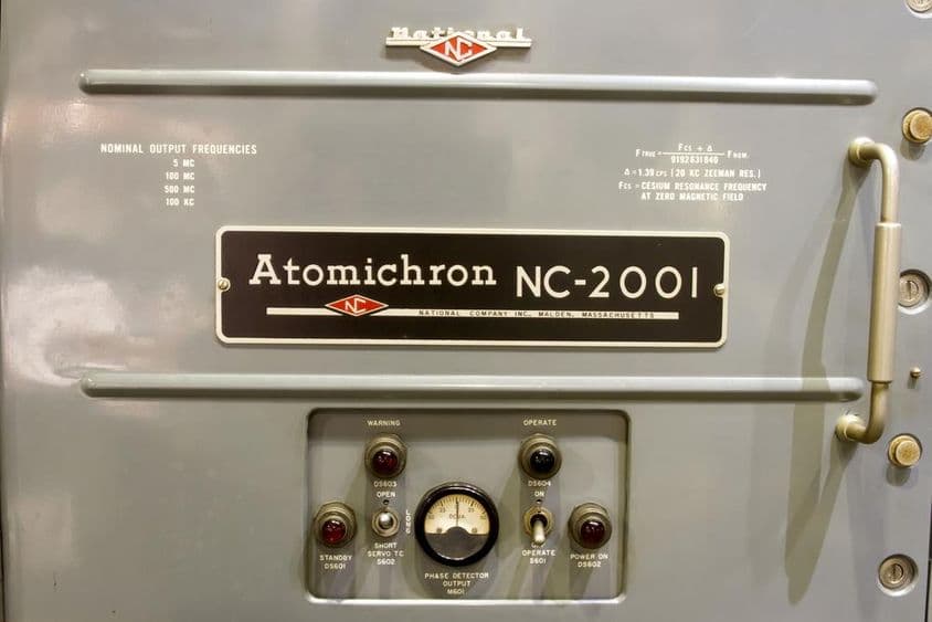 Dargestellt ist das Bedienfeld einer alten Atomuhr, zusammen mit ihrem Namen auf einem kleinen Schild.