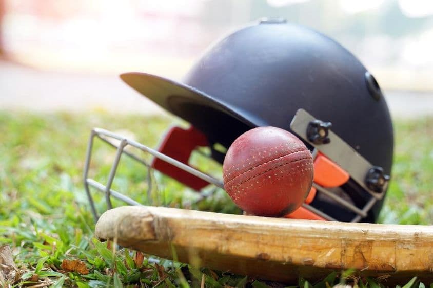 Ein Cricket-Schläger mit einem Ball darüber und dahinter, ein Cricket-Helm ist auf einem Grasfeld sichtbar.