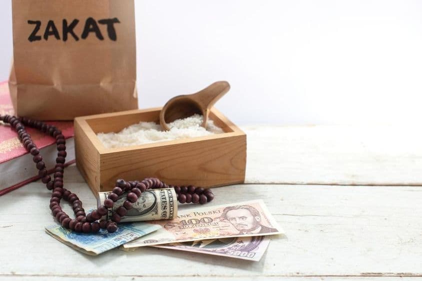 Mit 'Zakat' beschriftete Tasche vor einer Box mit Reis, Perlen, Dollars und anderen Währungen.