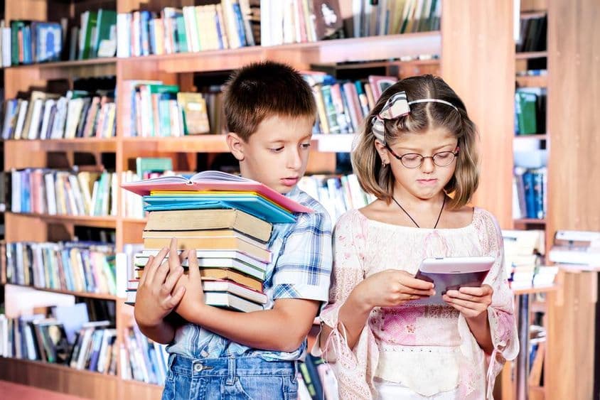 Zwei Kinder in der Bibliothek, eines liest ein E-Book, das andere trägt viele Bücher in den Händen.