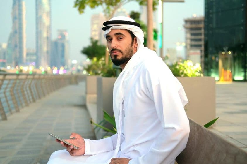 Emirati-Bürger sitzt auf einer Bank, hält ein Telefon, Wolkenkratzer im Hintergrund.