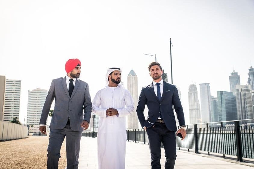 Ein Inder, ein Emirati und ein Europäer spazieren entlang des Kanals, Wolkenkratzer im Hintergrund.