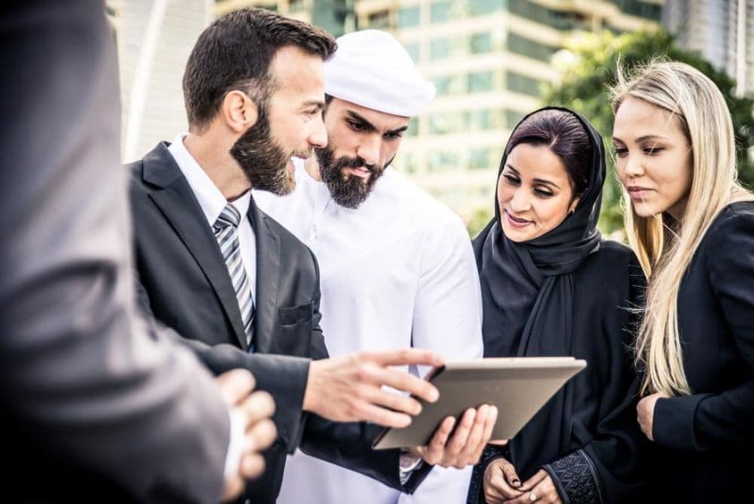 Ein europäischer und ein emiratischer Mann betrachten ein Tablet, flankiert von zwei Frauen, einer blonden und einer emiratischen, die zuschauen.