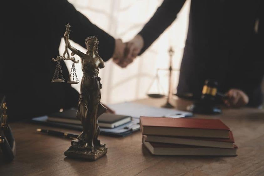 Ein Anwalt verhandelt mit einem Klienten, eine Justitia-Statue ist auf dem Tisch sichtbar.