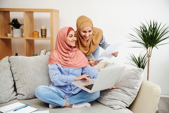 Zwei Emirati-Frauen überprüfen einen Vertrag und ein Notizbuch, eine hält ein Telefon, sitzen auf einem Sofa.