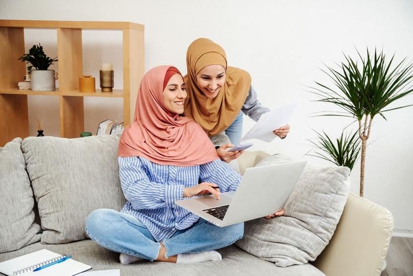 Zwei Emirati-Frauen überprüfen einen Vertrag und ein Notizbuch, eine hält ein Telefon, sitzen auf einem Sofa.