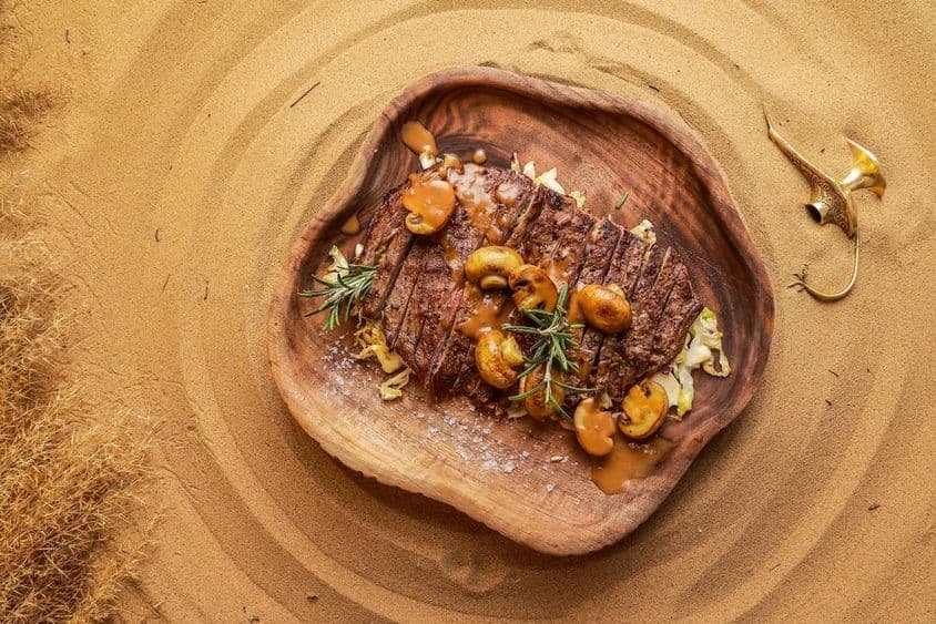 Steak serviert mit Pilzen auf einem Holzteller im Wüstensand, mit einer erloschenen Laterne daneben.