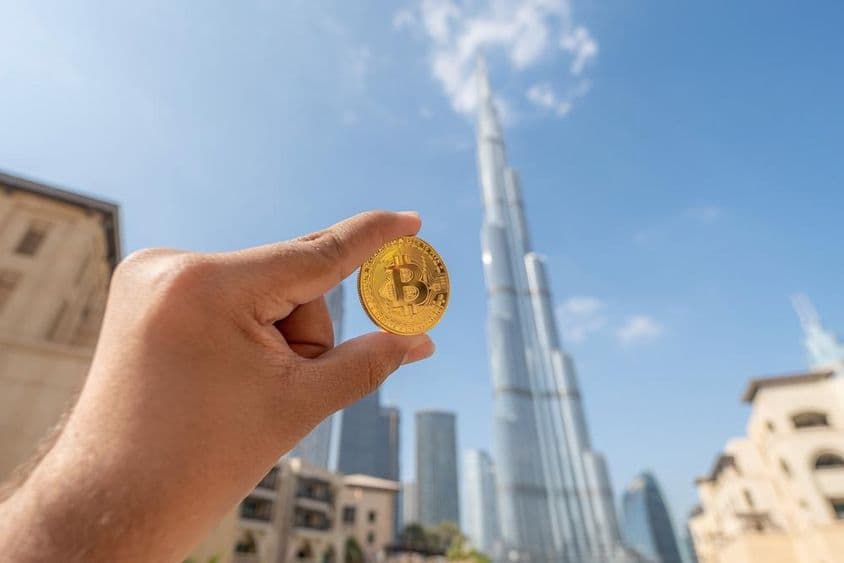 Person hält eine goldene Bitcoin-Münze, schaut in den Himmel, mit dem Burj Khalifa im Hintergrund.