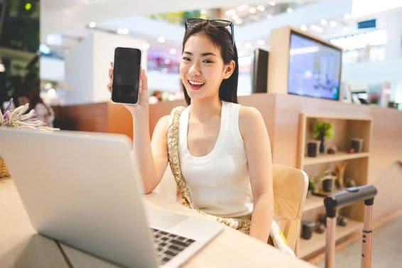 Gen Z-Mädchen arbeitet, hält ein Telefon, mit einem Notizbuch vor sich und einem Rollkoffergriff neben ihr.