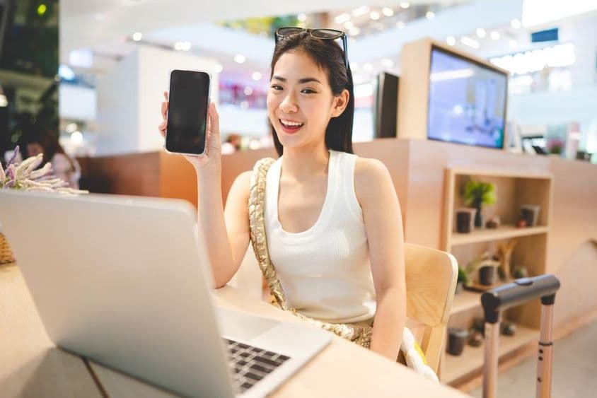 Gen Z-Mädchen arbeitet, hält ein Telefon, mit einem Notizbuch vor sich und einem Rollkoffergriff neben ihr.