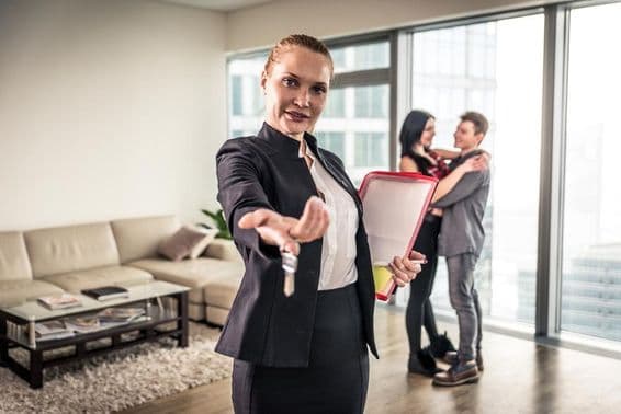 Eine weibliche Immobilienmaklerin winkt, ein Paar umarmt sich im Hintergrund.