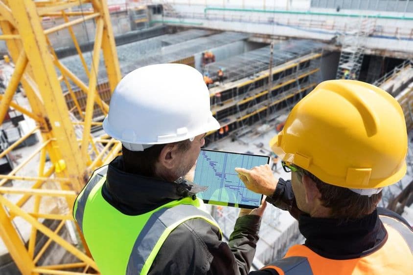 Zwei Personen mit Helmen auf einem Kran auf einer Baustelle, die Pläne auf einem Tablet überprüfen, mit der Baustelle vor ihnen.