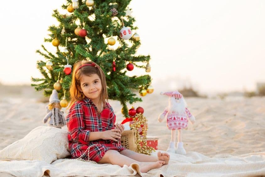 Fröhliches, lächelndes kleines Mädchen, das unter einem Weihnachtsbaum am Strand sitzt.