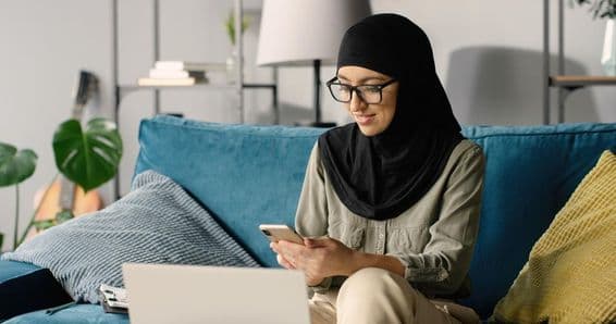 Arabisches Mädchen im Hijab arbeitet zu Hause mit Smartphone und Laptop auf der Couch.