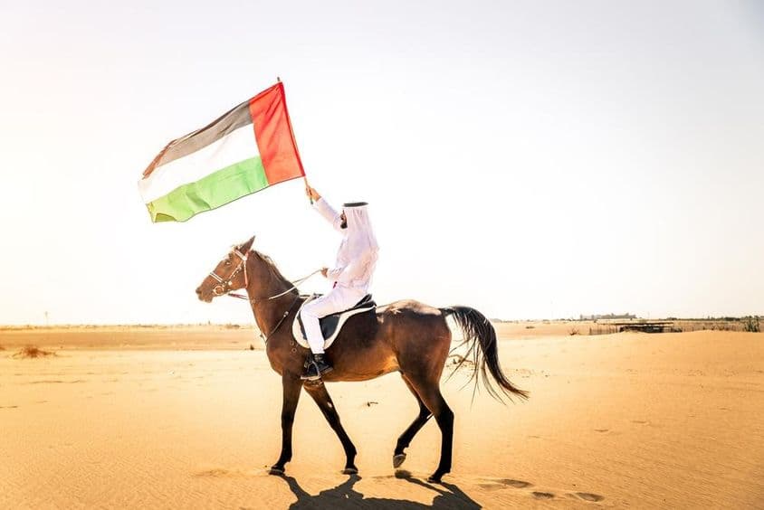 Mann aus dem Nahen Osten in traditioneller Emirati-Kleidung reitet ein arabisches Pferd in der Wüste Dubais.