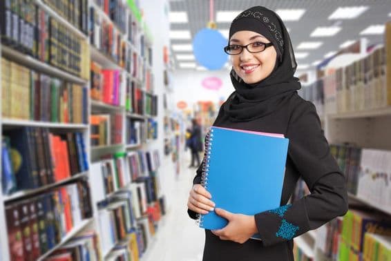 Arabischer Student steht in der Bibliothek mit Notizbuch in der Hand.