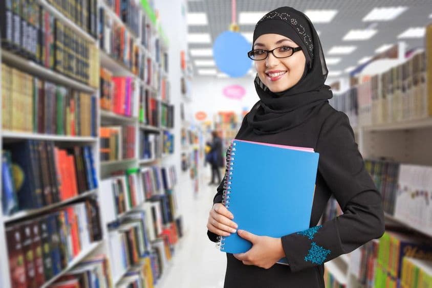 Arabischer Student steht in der Bibliothek mit Notizbuch in der Hand.