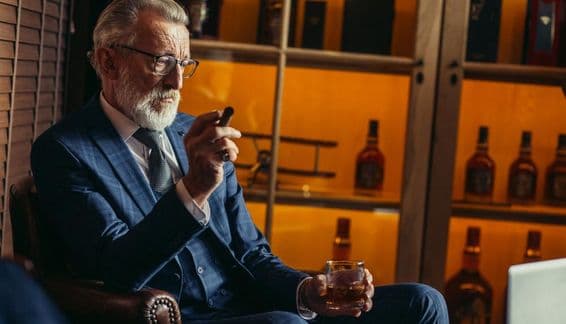 Älterer pensionierter Mann nutzt einen Laptop, raucht, hält teuren Alkohol in der Hand.