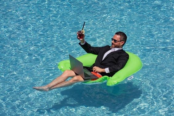 Erfolgreicher Geschäftsmann im Anzug schwebt im Poolwasser, trinkt einen Sommercocktail und benutzt einen Laptop im Pool.