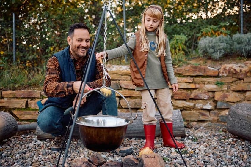 Kleines Mädchen hilft ihrem Vater, im Herbst in der Wildnis über einem Lagerfeuer zu kochen.