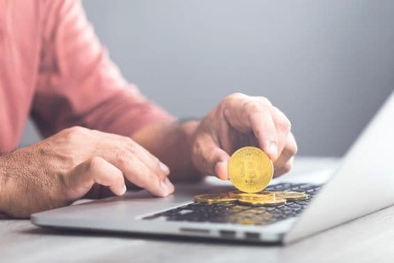 Ein Mann hält ein goldenes Bitcoin, während er einen Laptop benutzt.