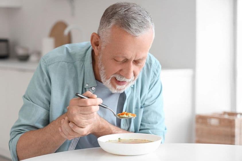Älterer Mann mit Parkinson isst an einem Küchentisch.