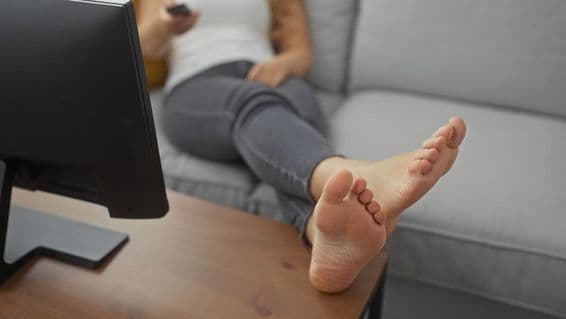 Eine junge Frau entspannt barfuß auf einem gemütlichen Wohnzimmer-Sofa mit einer Fernbedienung in der Hand.