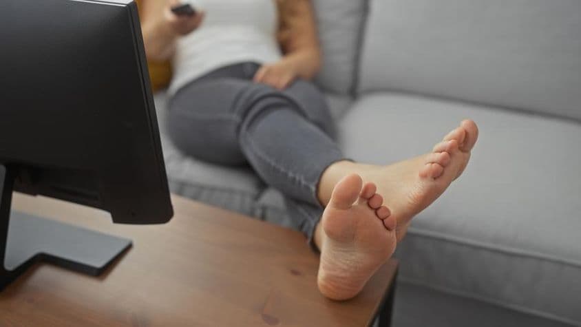 Eine junge Frau entspannt barfuß auf einem gemütlichen Wohnzimmer-Sofa mit einer Fernbedienung in der Hand.