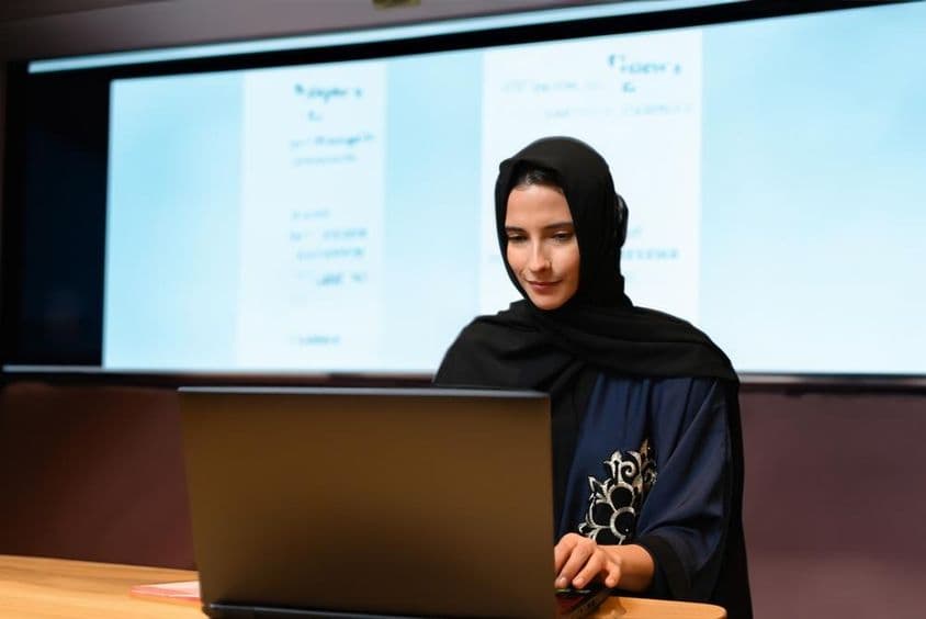 Lehrerin mit Abaya und Hijab vor einer Projektorleinwand in einem modernen Klassenzimmer mit digitalem Bildschirm.