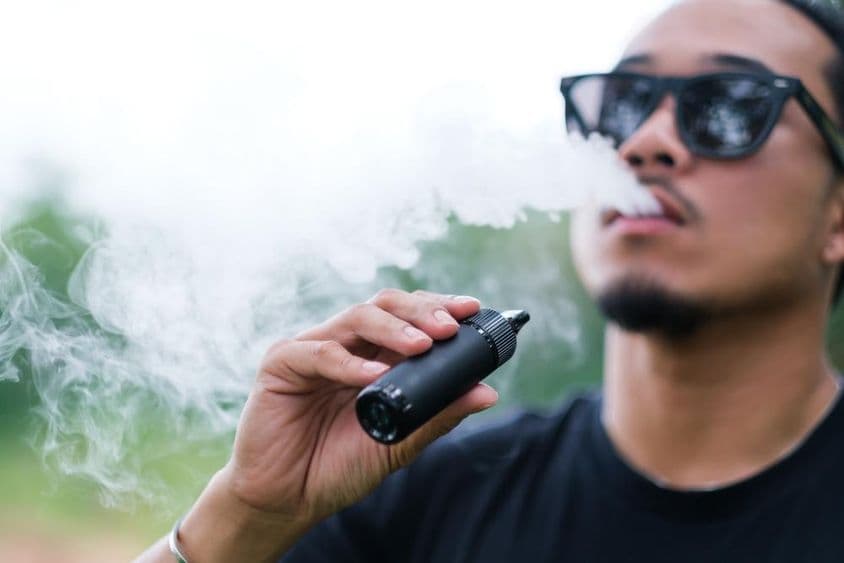 Asiatischer Mann raucht mit einer Vaping-E-Zigarette, stößt Rauch aus seinem Mund aus und genießt draußen im Sommer.