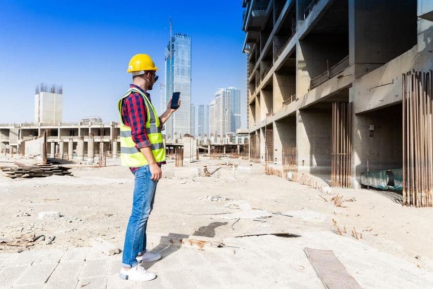 Mann benutzt Smartphone an einer Baustelle in Dubai mit Stadtsilhouette im Hintergrund.