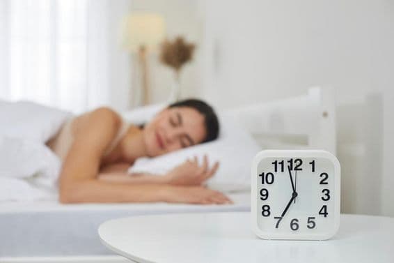 Wecker auf Nachttisch im Schlafzimmer, mit einer jungen Frau, die im Hintergrund schläft.