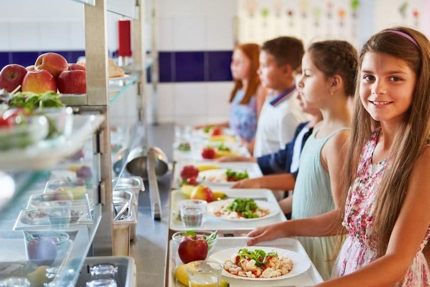 Lächelndes Mädchen mit Tablett, steht mit Freunden in der Warteschlange während der Mittagspause in der Schulkantine.