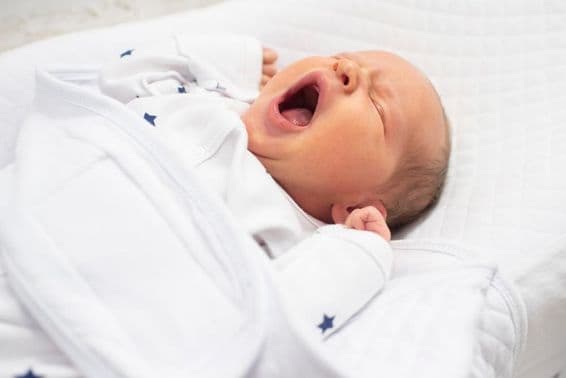 Ein einwöchiger Baby-Junge gähnt auf einem weißen Bett.