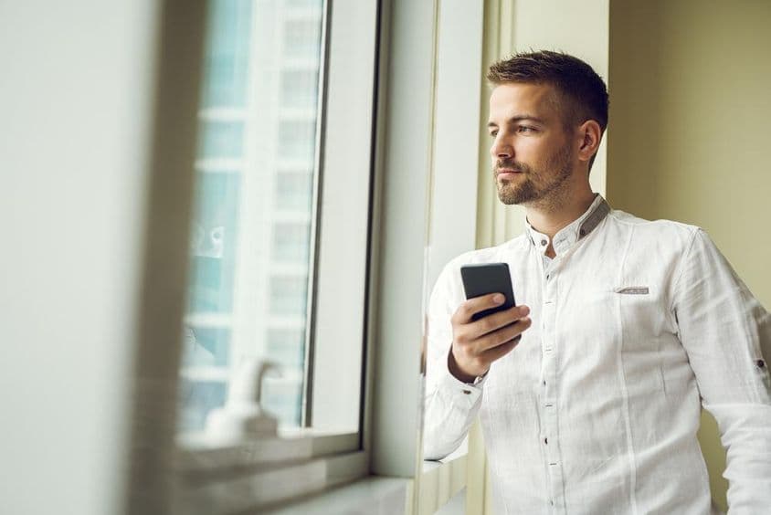 Geschäftsmann steht am Fenster, hält ein Smartphone.