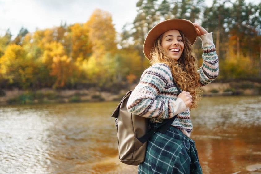 Weibliche Reisende mit Rucksack und Mütze genießt eine Herbstwanderung entlang eines Flusses.