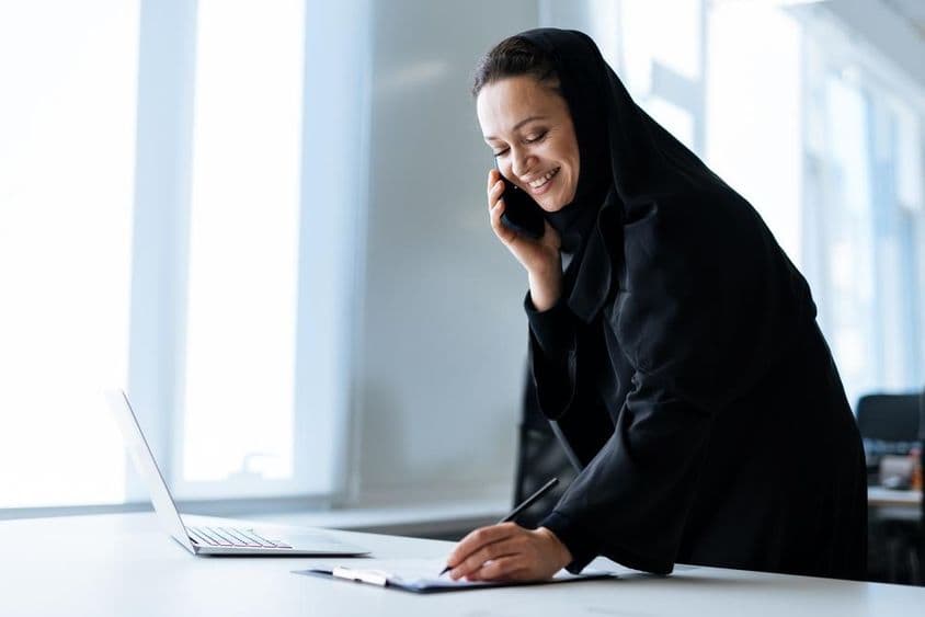 Eine schöne Frau in einer Abaya, die an ihrem Computer arbeitet, während sie telefoniert.