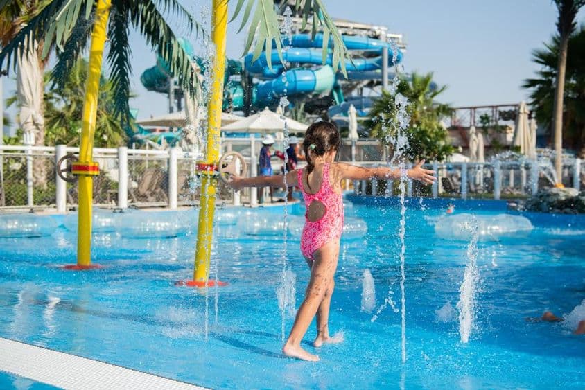 Kleines Mädchen spielt in einem Schwimmbecken mit Fontänen in einem Wasserpark.
