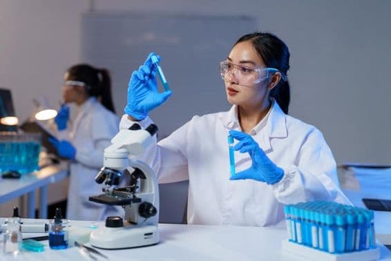 Asiatische Wissenschaftlerin im Laborkittel und Handschuhen, die zwei Reagenzgläser vergleicht.