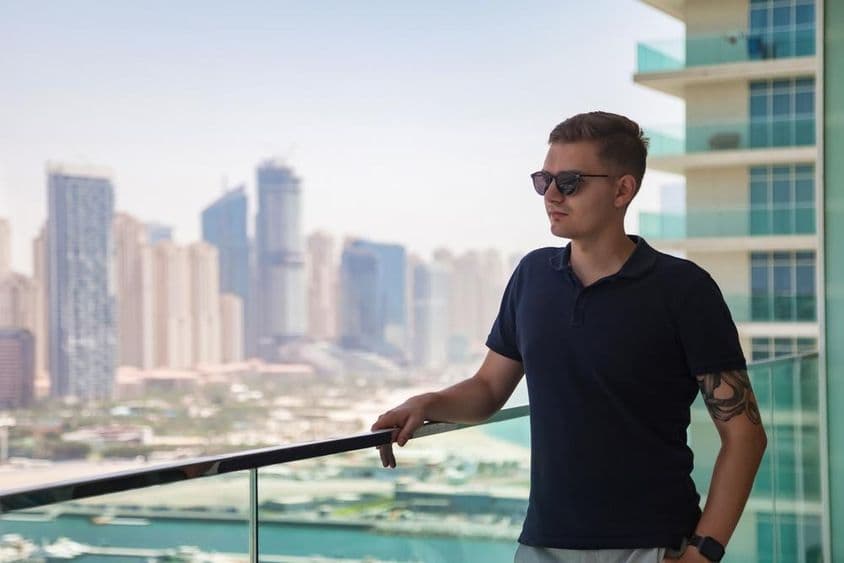 Ein junger Mann mit Sonnenbrille steht auf einem Balkon eines Wolkenkratzers mit Blick auf Dubai.