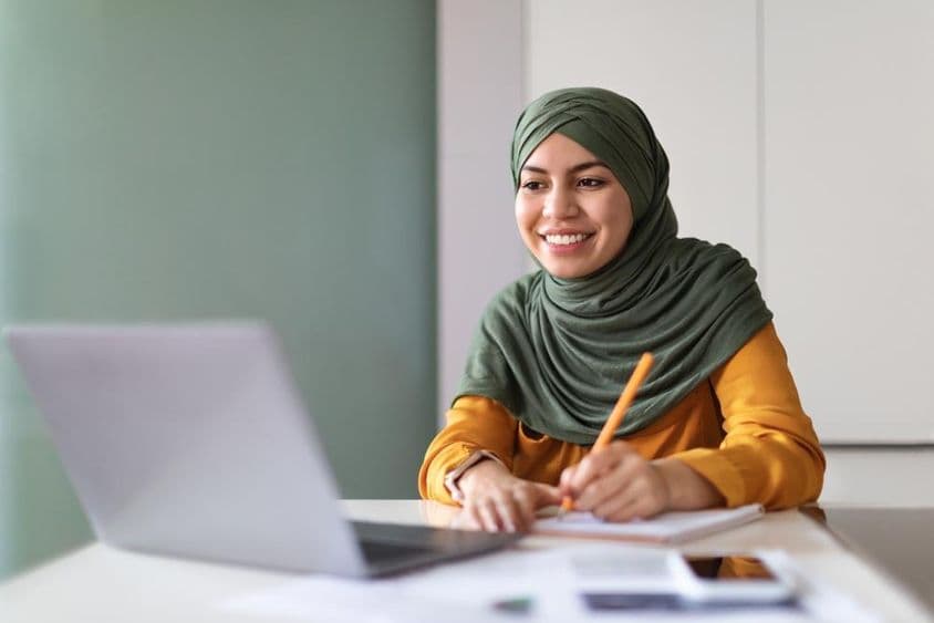 Junge muslimische Frau, die zu Hause an einem Webinar auf einem Laptop teilnimmt und Notizen macht.
