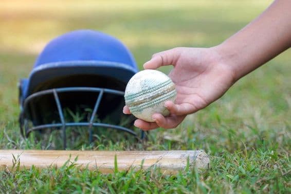 Die Hand eines Spielers hebt einen Cricketball aus anderen auf dem Gras liegenden Cricket-Ausrüstungen auf.