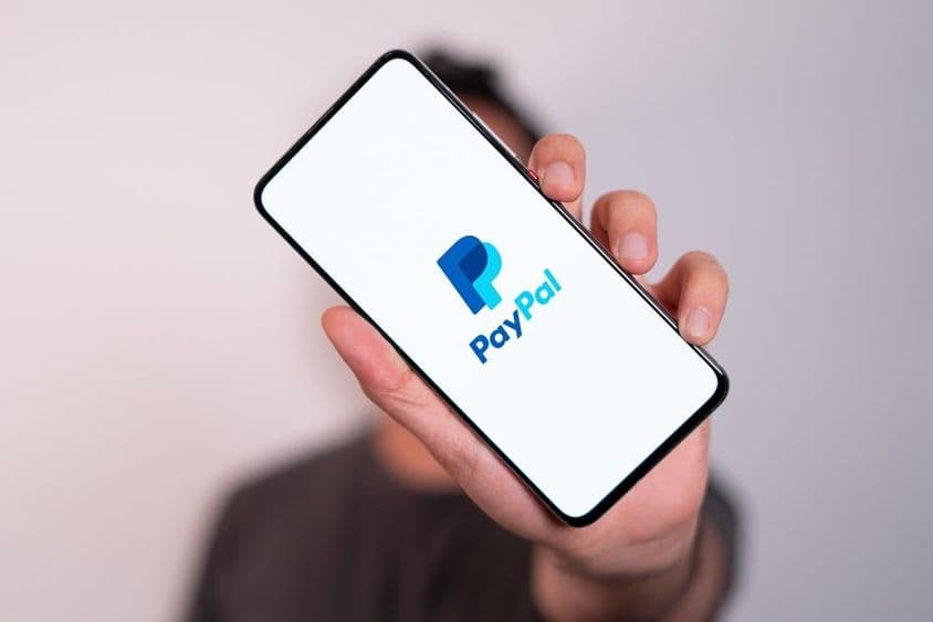 Ein Mann hält ein Telefon mit sichtbar auf dem Bildschirm das PayPal-Logo.